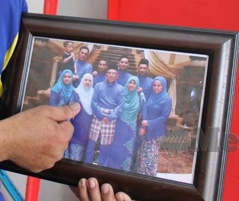 Norhaliza Hussin poses next to her husband and family members in a professionally-shot family portrait. Her husband holds a framed copy of the portrait and points at her.