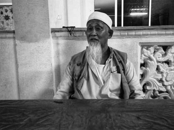 Ahmad Badarddin Mat Ripin, 64. An elderly man with long white beard. He is wearing a kopiah, a jubah (robe) and a vest on top of the robe. He is sitting at a table outside a building, which looks like a mosque.