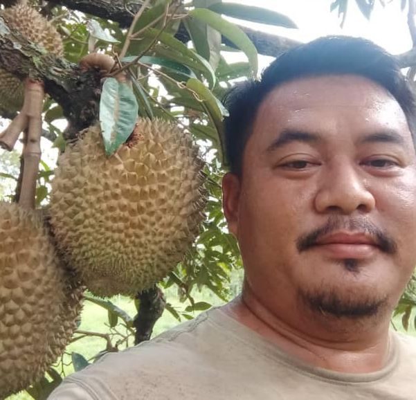 Ghani Jojo poses in front of durian fruits hanging from a tree. He has side swept short hair, a mustache and goatee.