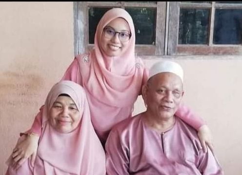 Md Nor bin Mohd with his wife and daughter. They are wearing matching pink baju melayu and baju kurung. His daughter has her arms around her parents. 