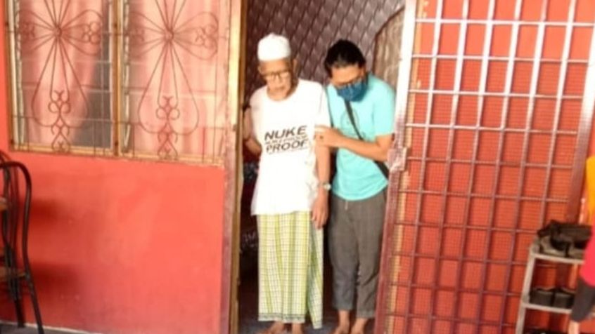 Misnon Ramli is assisted by his son out of the home. He is wearing a kopiah T-shit and sarong. The photo was taken before they left for the clinic, and minutes before he died en route.