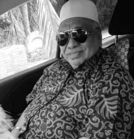 Mohd Saidi bin Mahat poses in a car. He is wearing aviator sunglasses with a white kopiah and a batik short sleeved shirt