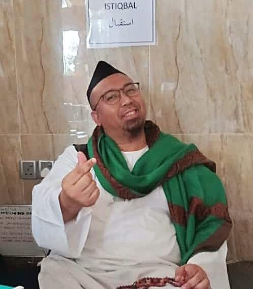 Nabil Azwar wears a half-rimmed glasses, a kopiah and jubah, and has a scarf around his shoulders. He is smiling and has a goatee. He gestures the 'mini love' sign wit his fingers.