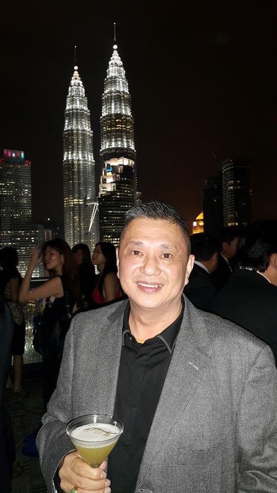 Chew Kar Yuen, 55, is holding a cocktail in a martini glass at a outdoor rooftop party at night. He is smiling. He is wearing a grey jacket with a black collared shirt with an open collar. In the background is the Petronas Twin Towers.