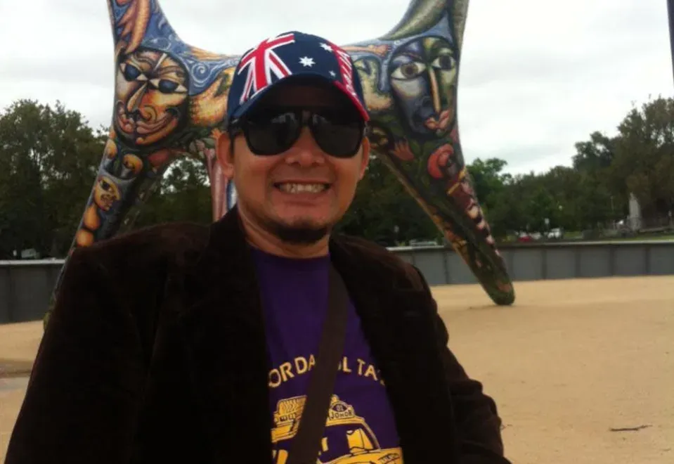 Azizan Khalid poses in front of a monument. He has tanned skin and a goatee. He is wearing a cap with the Australian flag, sunglasses, a jacket, a T-shirt and a sling bag. He is smiling.