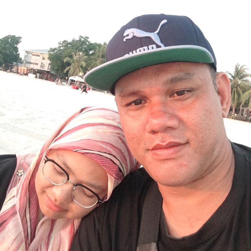 Abdul Hadi Mohd Yunus and his wife on a beach. His wife is leaning on his shoulder. He is wearing a cap and a black T-shirt.