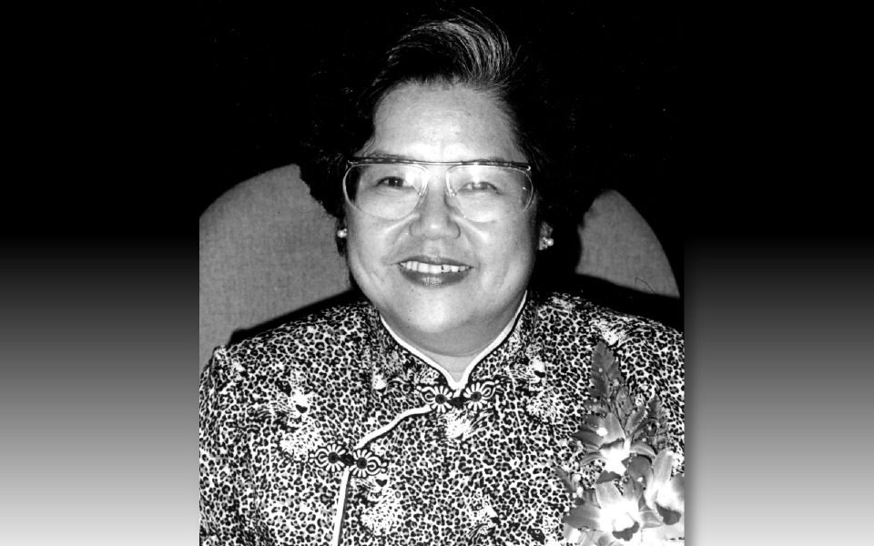 Kee Phaik Cheen smiling to the camera. She has a grey streak in her hair, wears wide framed glasses and a cheongsam.