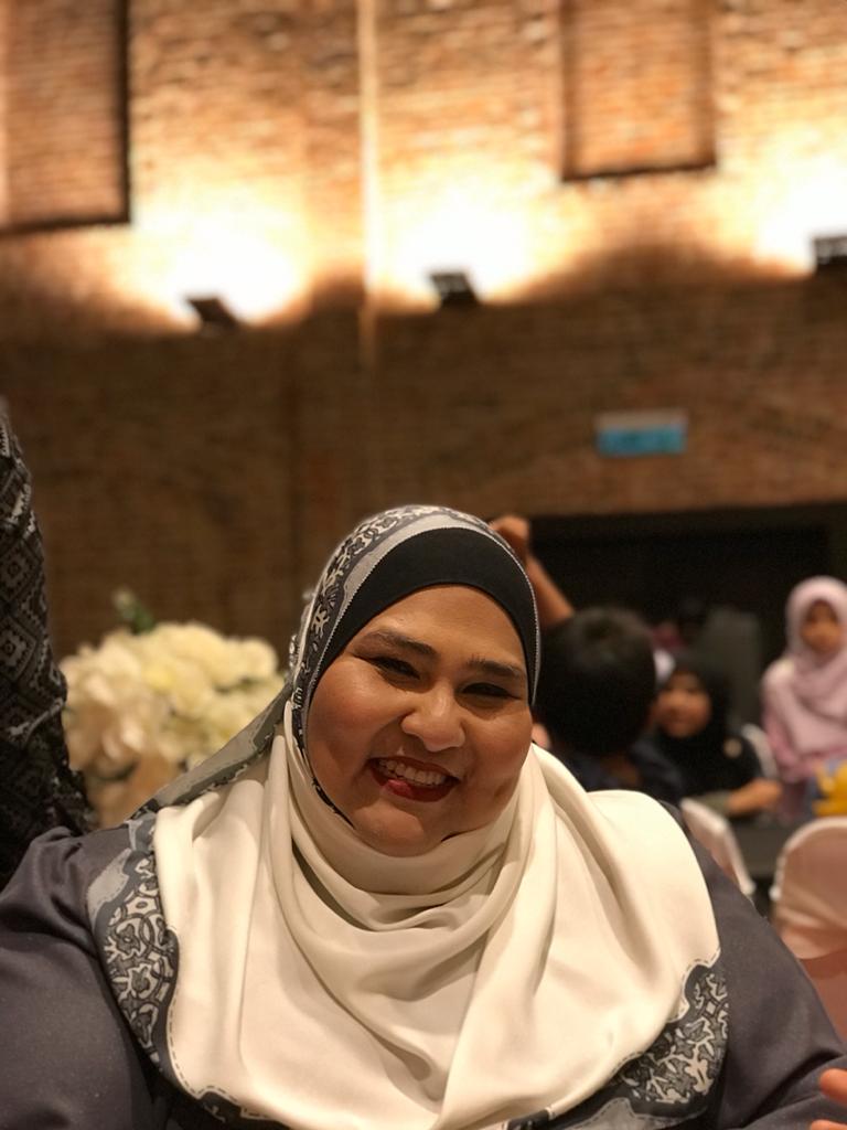 Sit Majmin Jalani smiles whiel attending a wedding. She is large woman with dimples and high cheekbones. She is pictured wearing red lipstick, a headscarf and baju kurung.