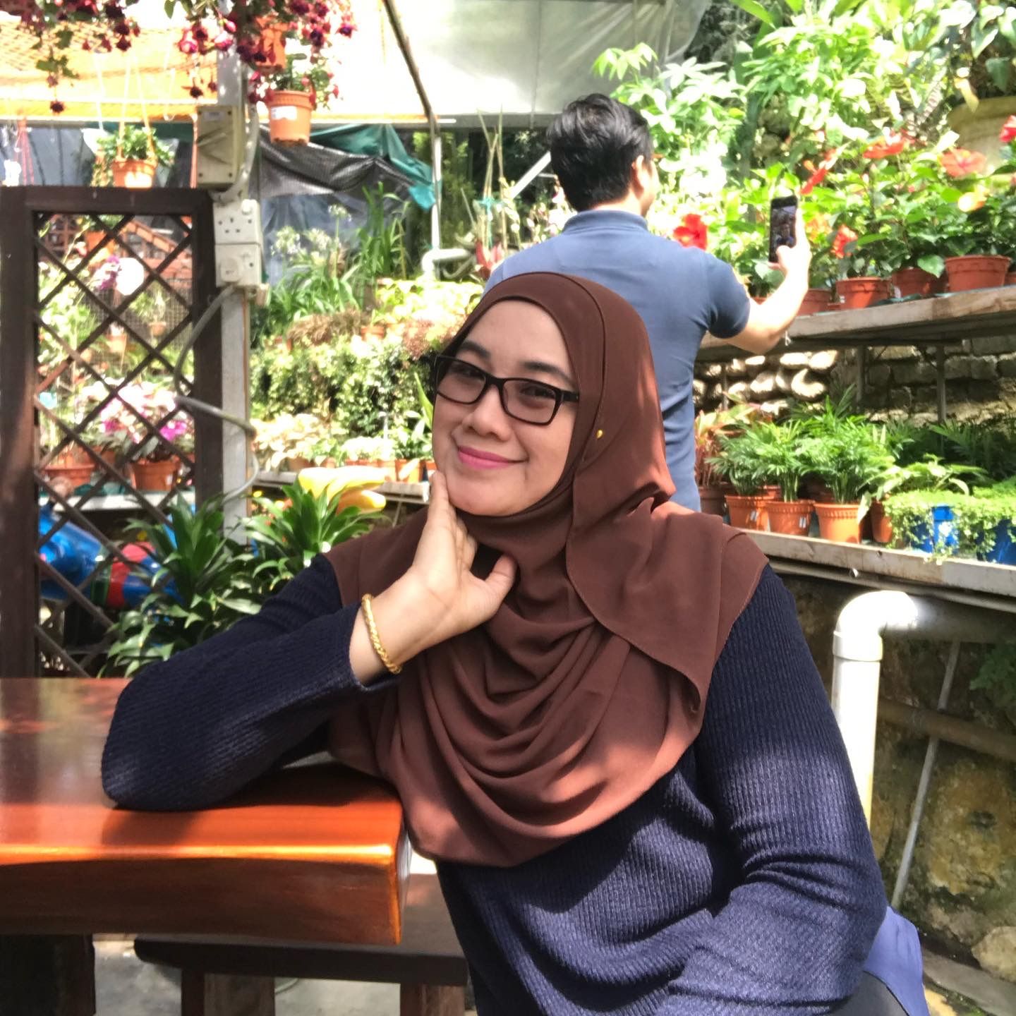 Hairiza binti Anuar Misbah is smiling. She is sitting at a nursery. She wears a brown headscarf, black top, glasses and a bracelet.
