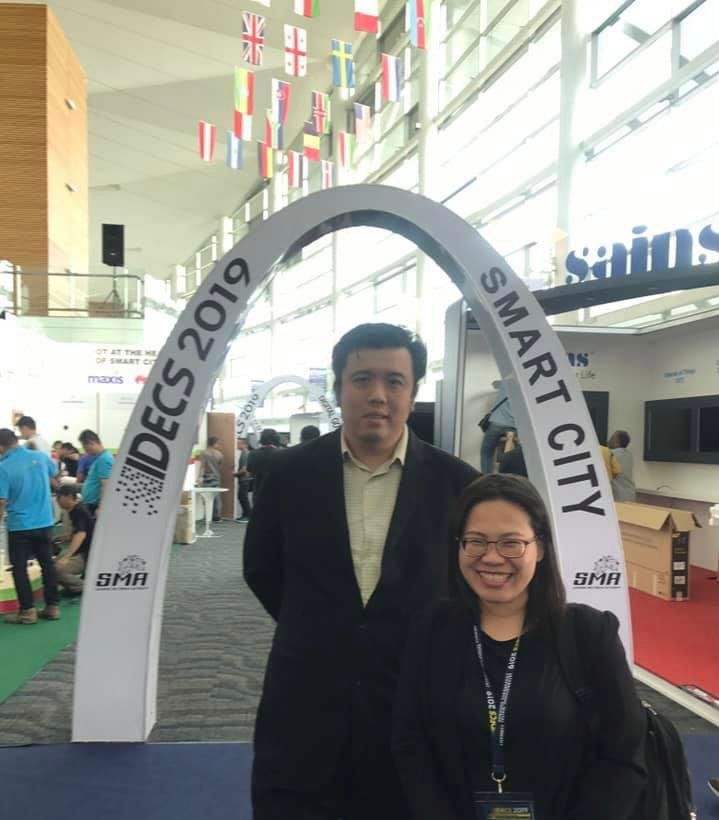 Lee Jun Choi, in a black jacket and cream shirt, stands behind colleague Emmy Dahliana, at the IDECS Smart City exhibition in 2019. He is tall, with short, black hair.