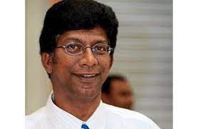V Sivalingam smiles in a candid shot. He has tanned skin, thick hair and wears rimless glasses. He is wearing a shirt and a tie.