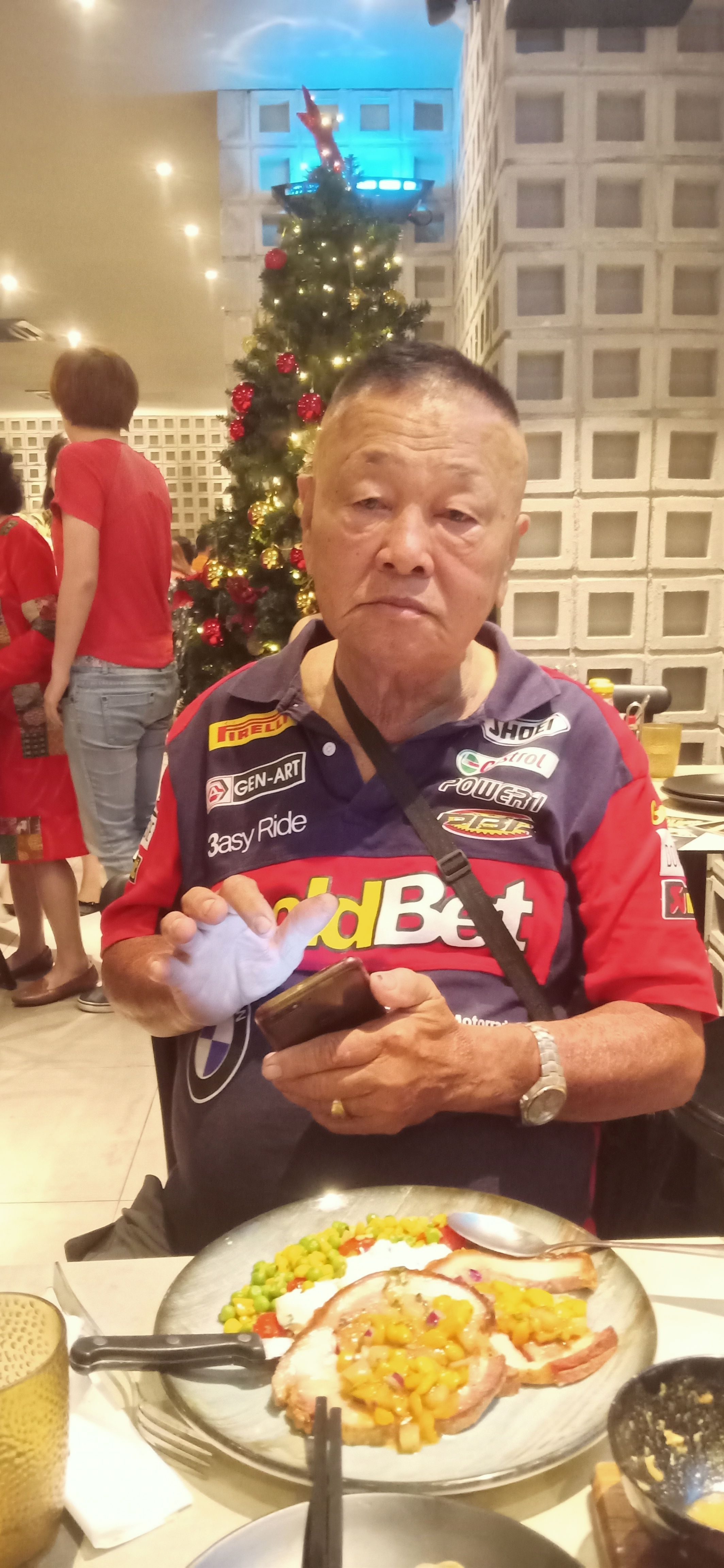 Lee Tam Sing holds his phone at the dinner table. He wears a collared T-shirt, a sling bag and a silver watch. He has short cropped hair. In the background is a Christmas tree.