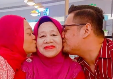 Noraini Othman is kissed by her children. She is wearing bright red lipstick which matches her red headscarf and blouse.